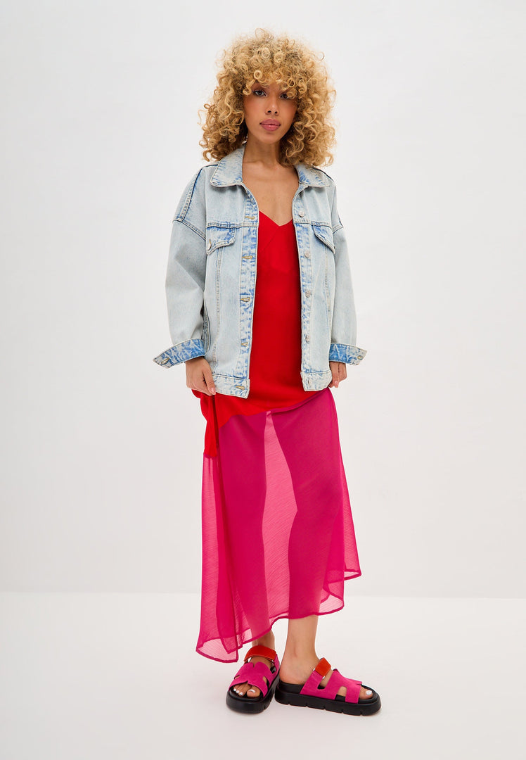 Fashionable woman in a red top and sheer pink skirt, styled with a denim jacket and pink platform mules, showcasing a chic and casual look.