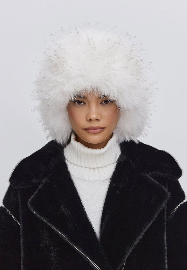 Woman wearing a stylish winter fashion hat, the Irina faux fur hat in black and white, paired with a black coat and white turtleneck.